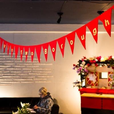 Red bunting that reads "Young Vic two buroughs" hangs from the ceiling of a room with a sofa and a small table covered in flowers
