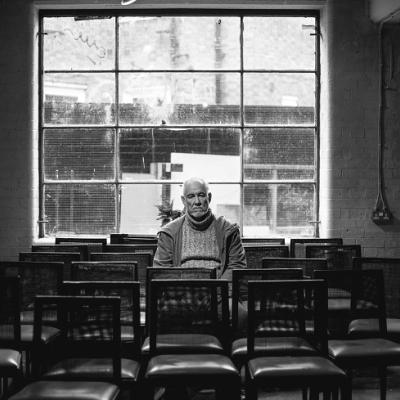 A room with a large window at the back full of empty chairs except for one elderly man wearing a jumper and jacket with a sad expression sat in the centre.