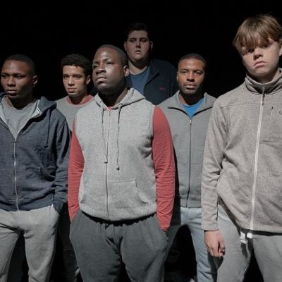 A group of 6 young people in tracksuits stand in a black space with serious expressions 