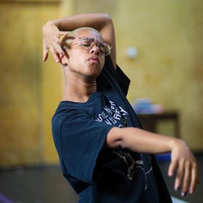 A person wearing a black t-shirt and glasses with short shaved blonde hair stands in a voguing pose with their eyes closed 