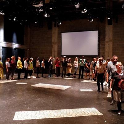 A large group of people walk in a semi-circle following each other in the Young Vic studio space with a white screen at the back and seated audience on either side of the room watching them