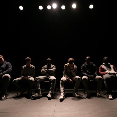 The group of six male actors sit backlit on their seats at the far end of the stage. We are not able to see any of their faces but their body language is negative, seemingly annoyed or put out © Leon Puplett
