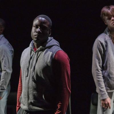 A man in a red and grey hoodie stands half in light with a solemn expression looking out to the audience as two other make performers stand either side of him a few feet behind him © Leon Puplett