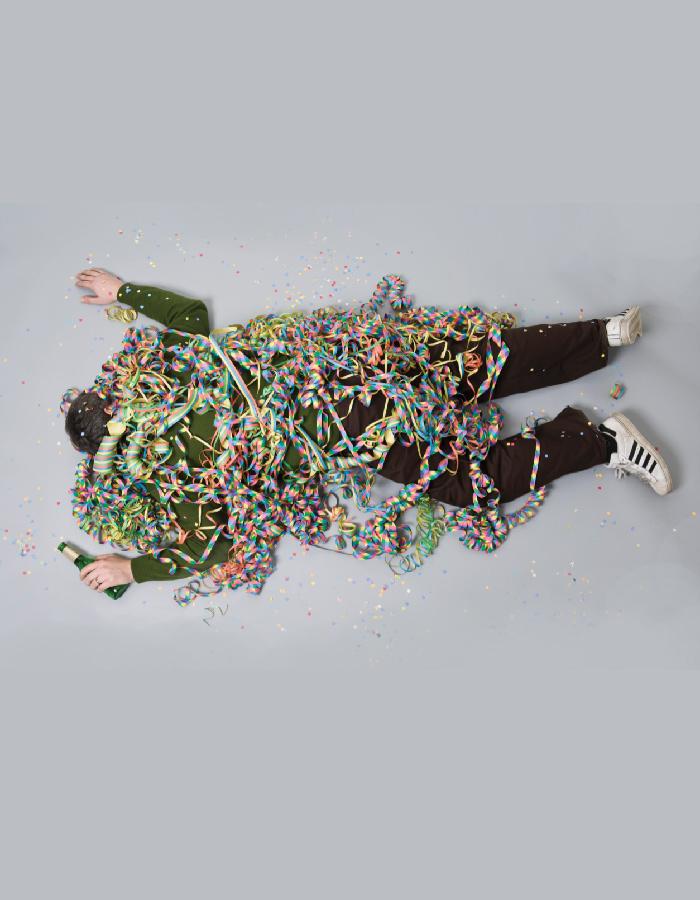 Man lies on a grey floor with beer bottle in hand, covered in confetti streamers