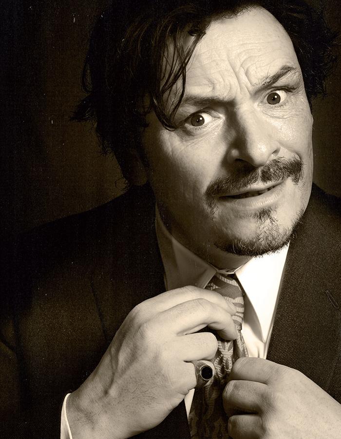 Julian Barratt looks quizzically at the camera while tightening his tie. 