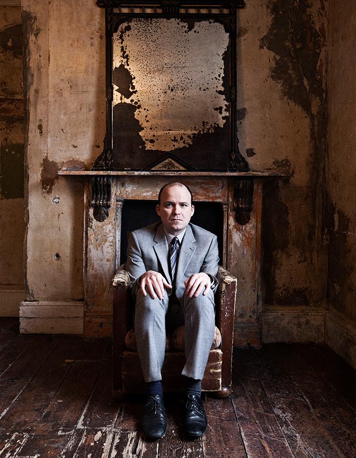 Rory Kinnear sits in a chair under a mirror in a derelict room 