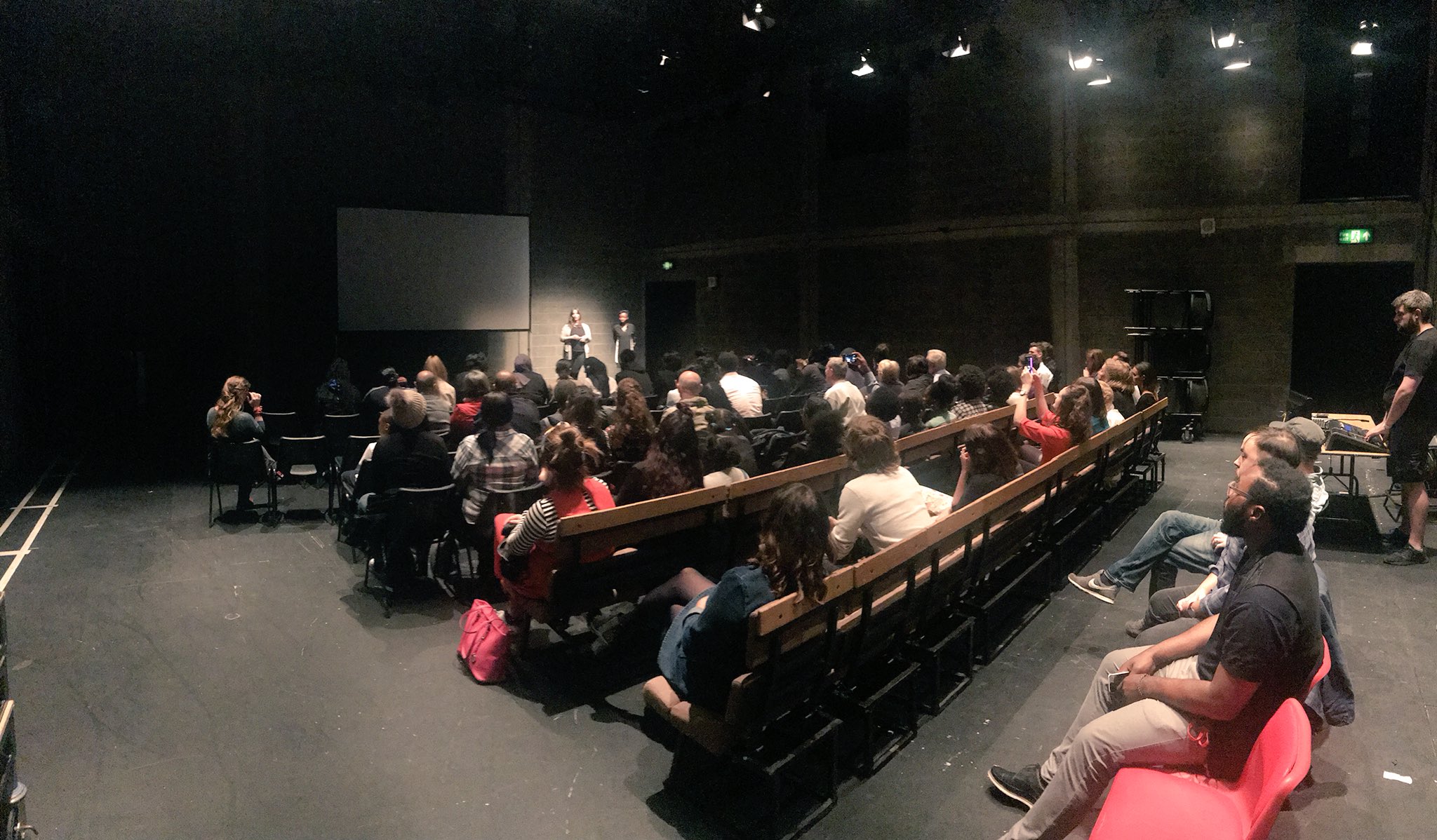 A busy audience of friends and family sit in rows in front of a projector screen ahead of the London Fable sharing in the YV's Maria theatre