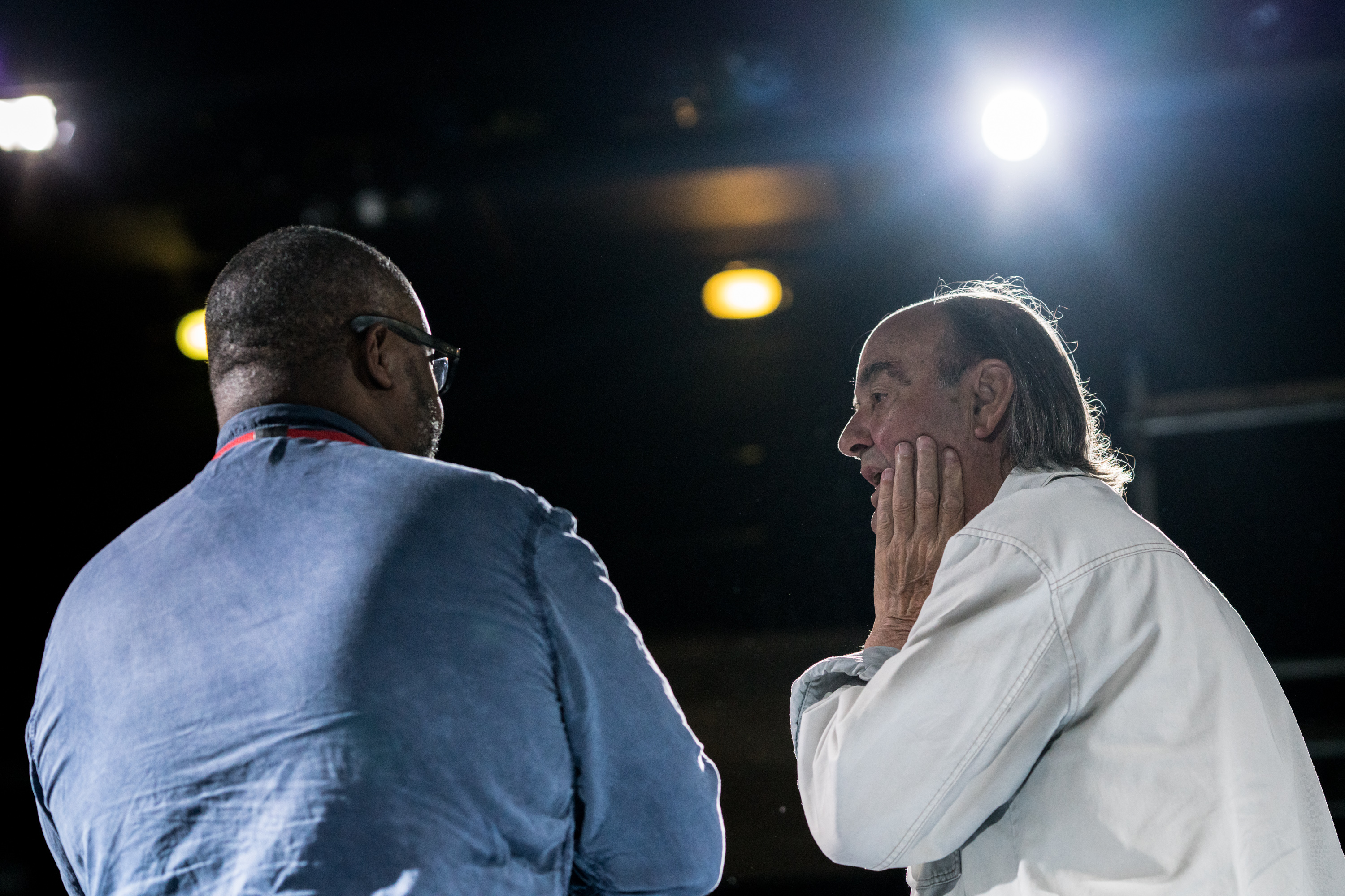 An actor from Life of Galileo is talking to an audience member, who listens with a look of rapt attention, his hand up to his mouth, his mouth open. The two men have their backs to us but their heads are turned inwards, towards each other, so that we can see their expressions. A stage light shines brightly in the background, out of focus.