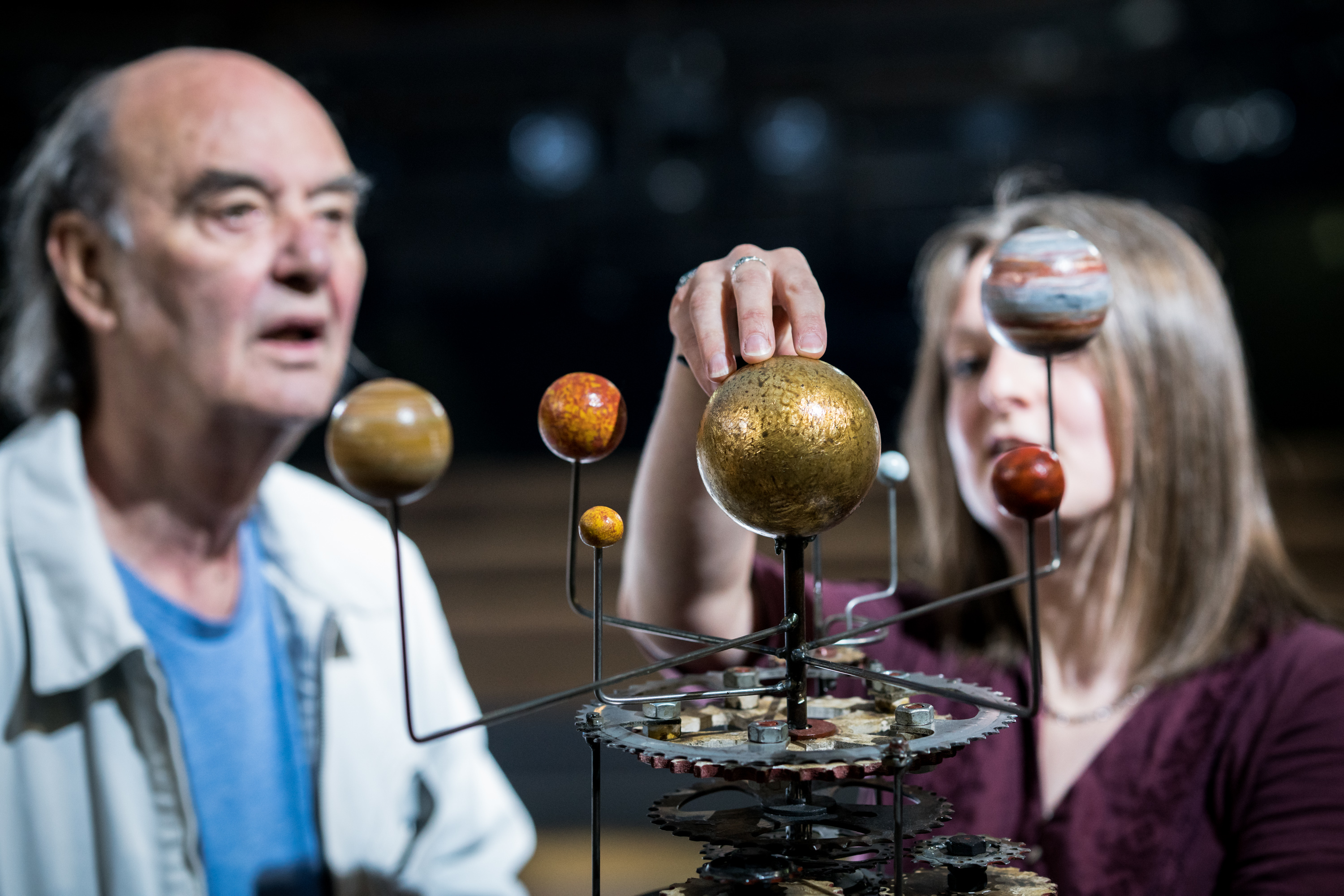 An audience member listens to the Life of Galileo stage manager describe a heliocentric model of the solar system prop from the show. The audience member and stage manager are in the background of the image with the focus on the heliocentric model of the solar system in the foreground with the stage manager’s hand placed on the sun.