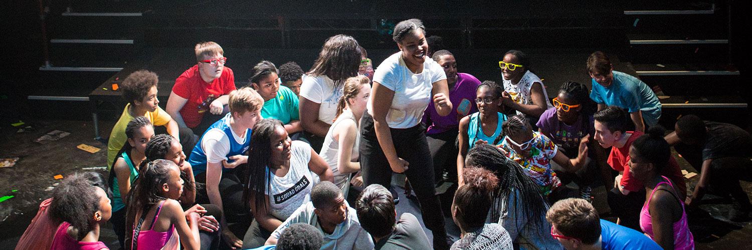 A single performer stands dancing amongst a group of school age performers who are kneeling in a circle around them. Production photo by Helen Murray of Taking Part's Stay Another Song schools production.