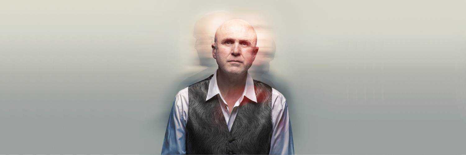 A head and shoulders shot of Tim Crouch wearing a white long sleeve shirt and a patterned gray vest. Bahind him are blurry shadows of his head facing the left and right. 