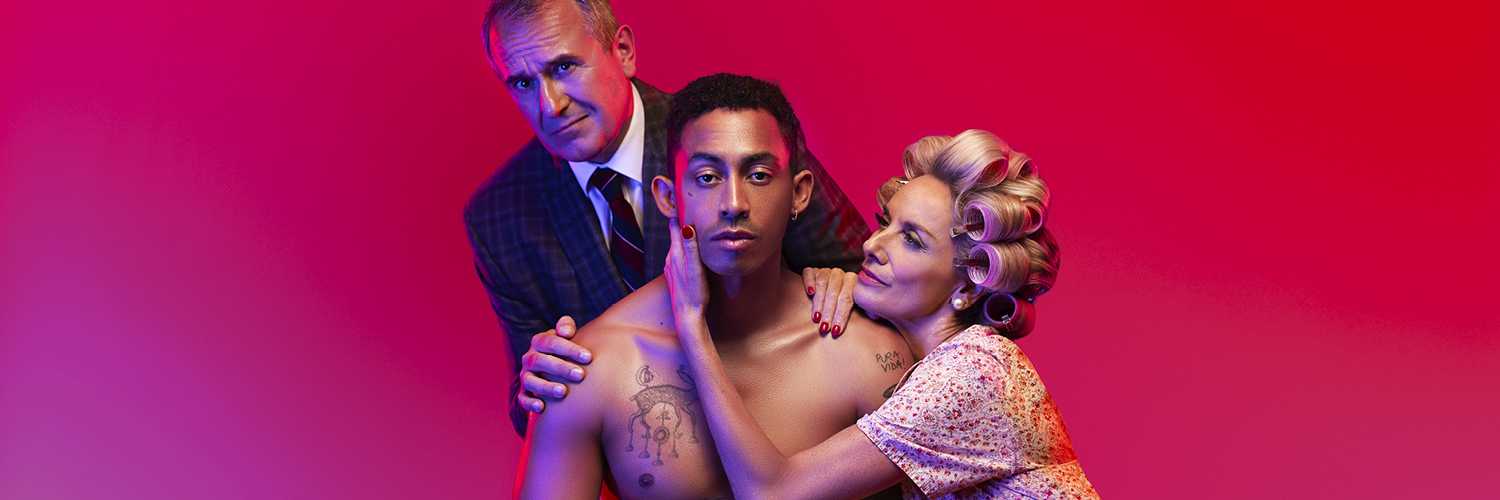 3 people pose together against a pink background. The person in the centre is sat on a chair wearing white underwear. A person kneels on the right with one hand cupping the face of the person in the middle. A person stands on left with both hands on the shoulder of the person in the centre. 