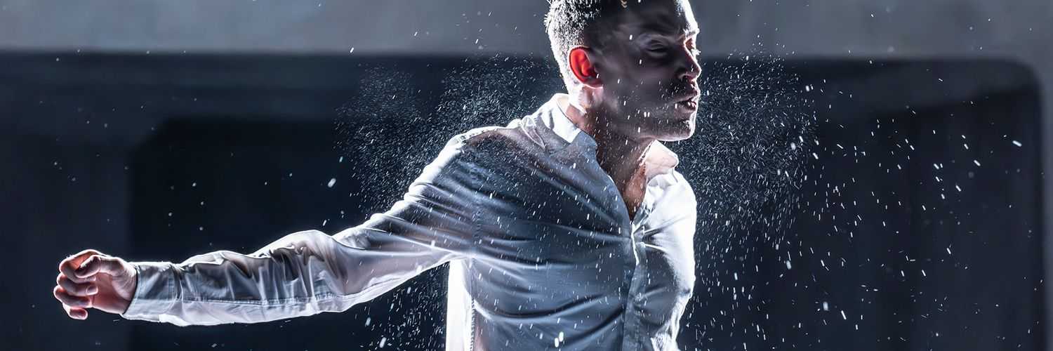 A man lit from behind, wearing a white shirt and blue jeans is standing on a stage. His arms are spread out to the side, and his eyes are closed. He is mid-action and there are sprays of water emanating from him