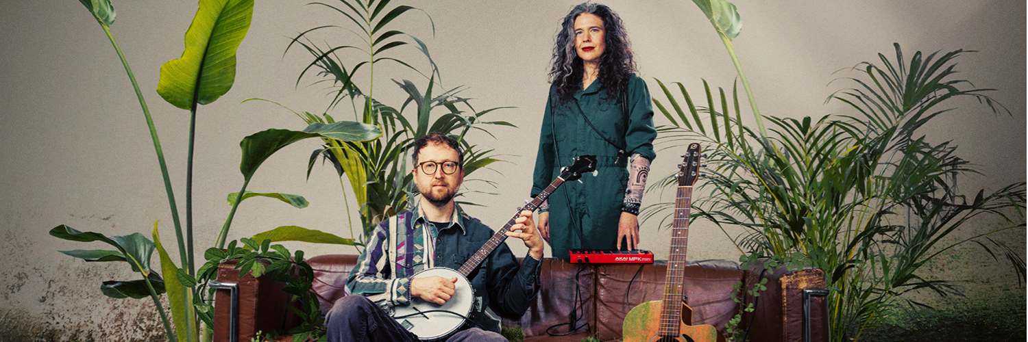 Against a background of green plants are a man and woman. The man sits on a sofa and is holding a banjo; the woman stands behind the sofa. Next to them is an orange guitar on a stand. 