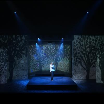 Irfan Shamji standing holding a baby, with projections on the wall in the background showing trees. Akhila Krishnan.