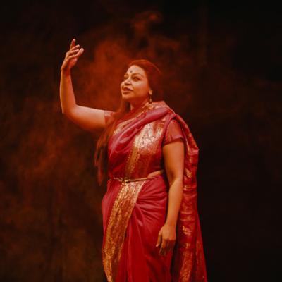 Ayesha Dharker standing with arms raised, wearing red sari with ornate gold pattern. Isha Shah.