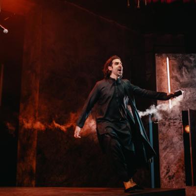 Scott Karim wearing a black kurta, running across the stage, holding a smoke flare. Isha Shah.