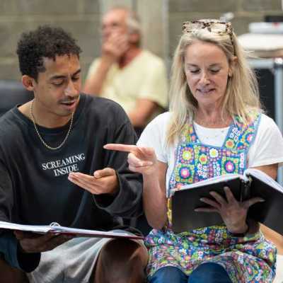 A young man with short, curly dark hair, wearing a long-sleeved black shirt, and a blonde woman wearing a colorful apron, both sit on a white chaise couch in a theatre rehearsal room. They perform a scene together, holding scripts