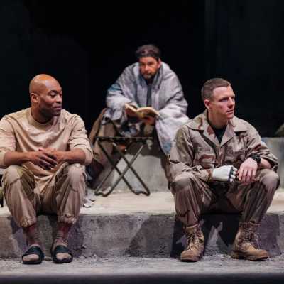 Three people sit on concrete steps. Two, in army uniform, sit on the front step. They aren't looking at each other but are in conversation. The third person has a blanket over their shoulders and is reading a book. He looks towards the two soldiers.