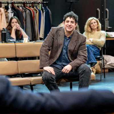 In the rehearsal room, an actor with short hair in brown blazer sitting on the bench facing anotehr actor with grey hair wearing a navy shirt back to the camera. Two actresses sitting behind him out of focus.