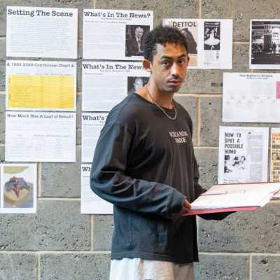 A young man wearing a long-sleeved black shirt stands in a theatre rehearsal room, in front of a wall of printed-out news articles
