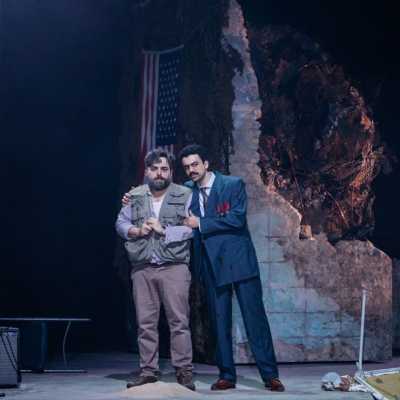 A man in a suit has his arm around a man in an army vest and civilian trousers. The man in the suit looks clean and confident, the other man looks scared. Behind them is a crumbling wall. The lighting is dark. 