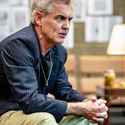 A close up shot of an actor with grey short hair wearing a navy blue shirt and a pair of green trouser sitting on the bench leaning forward with his hands crossed, elbows resting on his knees
