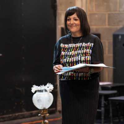 A woman with shoulder-length dark hair stands holding a script in a theatre rehearsal room. She stands behind an old-fashioned side table with a lamp, a chest, and a shell sitting on top