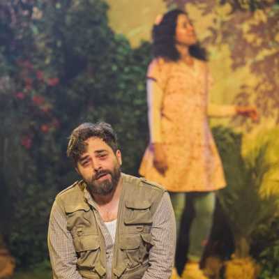 A man in an army vest and civilian trousers kneels at the front of the stage. Behind him, slightly blurred, is a young girl in a yellow dress with jeans underneath. She is looking at a beautiful garden