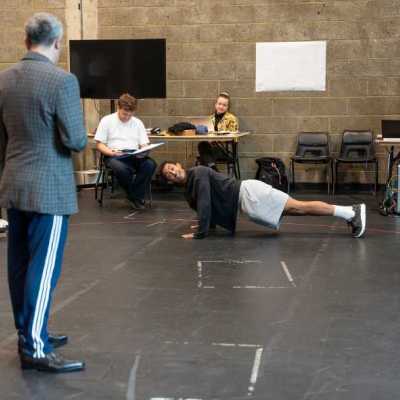 A man with short grey hair and a young man wearing a black shirt act in a scene together. The young man does push ups on the floor of the theatre rehearsal room. Several people watch from a table in the back of the room