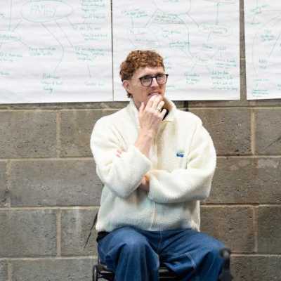 A person with short, curly red hair, and wearing a fluffy white jacket, sits in a theatre rehearsal room watching the actors work out of frame