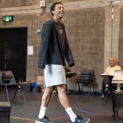 A young man wearing a black long-sleeved shirt walks across a theatre rehearsal room, laughing