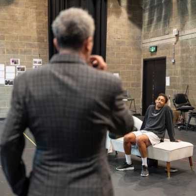 A man with short, grey hair wearing a grey suit stands with his back to the camera, performing a scene with a young man wearing a long-sleeved black shirt sits back casually on a white chaise couch. They are in a theatre rehearsal room