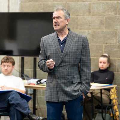 A man with short, grey hair rehearses for a play in a theatre rehearsal room. He wears a patterned grey suit and holds a prop cigarette
