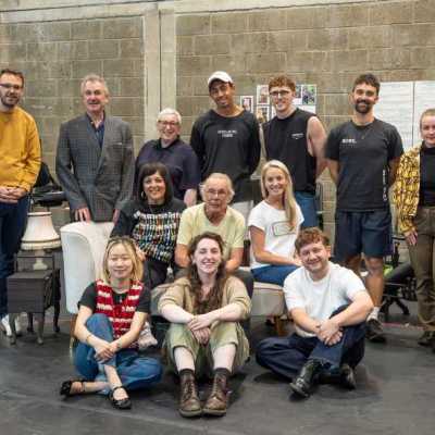 A group of actors and creatives pose smiling for a group photo in a theatre rehearsal room