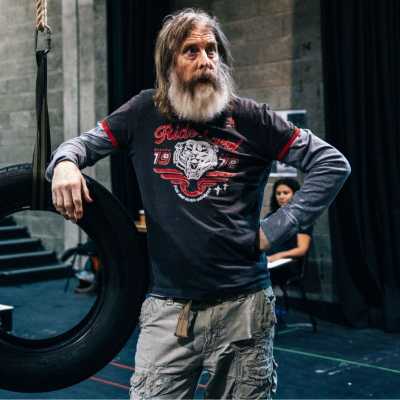 A person with a long beard wearing layered shirts leans against a tire swing in a theatre rehearsal room. 