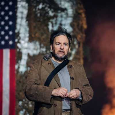 A man in a heavy overcoat stands in front of an American flag. Next to it, a warm light gives the impression of a burning fire. 