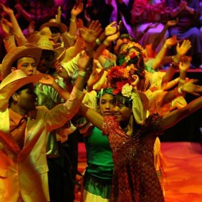 A line of men, in cowboy hats, and a line of women, with flower garlands in their hair, stood next to each other with their arms in the air. The two people in the front are smiling at each other