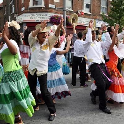 A group of people, in colourful dresses and cowboy hats are dancing in the street with their arms in the air. Some are holding hats aloft