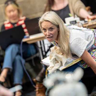 In a theatre rehearsal room, a blonde woman wearing a colourful apron sits on a bench, leaning over to grab something beneath her