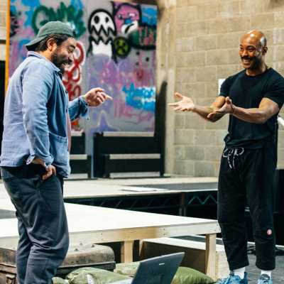 A person wearing black trousers and a black shirt and a person wearing a denim shirt and a baseball cap stand smiling and facing each other in a theatre rehearsal room. 
