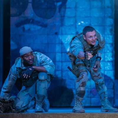 two soldiers stay low in front of a crumbling wall with graffiti on it. One soldier is kneeling and one standing but crouching forward. They are both looking to the left. 