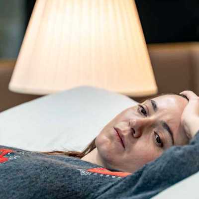 A closeup shot of a person in gey jumper lying on bed with one of her hands placing on her forehead. There is a lamp in the background 
