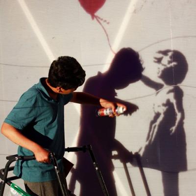 A young person holding a walking frame spray painting on a wall. On the wall, their shadow overlaps with the shadow of a little girl holding a red balloon.
