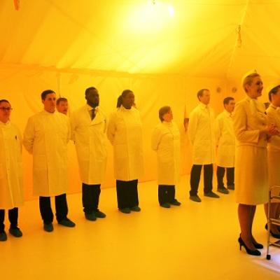 A line of people in white labcoats stand behind two people in white skirt suits and nurses caps in a tent space illuminated with yellow light. A person's head peers through the back of the tent.