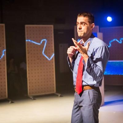 A person in a blue button shirt and red tie holds a bag over one shoulder and gestures with one arm in front of white panels, a large blue H and a small E