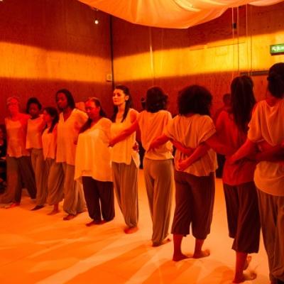 A group of people stand in a line with arms around each others' waists, with half facing one way and half facing the other, inside a tent lit with orange light