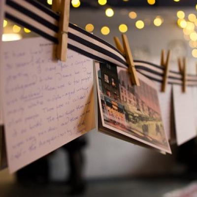 A striped clothesline with multiple wooden clothespegs and two postcards, one showing the front of the Young Vic and one with handwriting, hangs across the ceiling of an out-of-focus room with fairylights visible in the background 