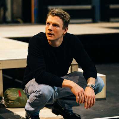 A person wearing jeans and a long-sleeved black shirt crouches on a platform in a theatre rehearsal room.