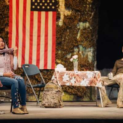 a woman in a headscarf and jeans sits at the front of the stage. A soldier sits near the back looking away from her. There is a basic table and plastic chairs and an American flag on the back wall. 